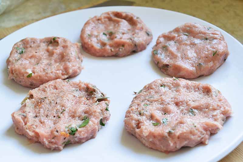 Horizontal image of ground turkey that has been mixed with herbs and shaped into five round patties, arranged in a circle on a white ceramic plate, on a beige speckled background.