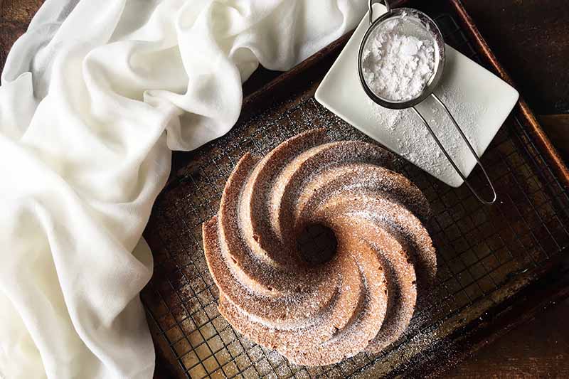 Horizontal image of a whole bundt covered in powdered sugar next to a strainer with more powdered sugar, all on a cooling rack.