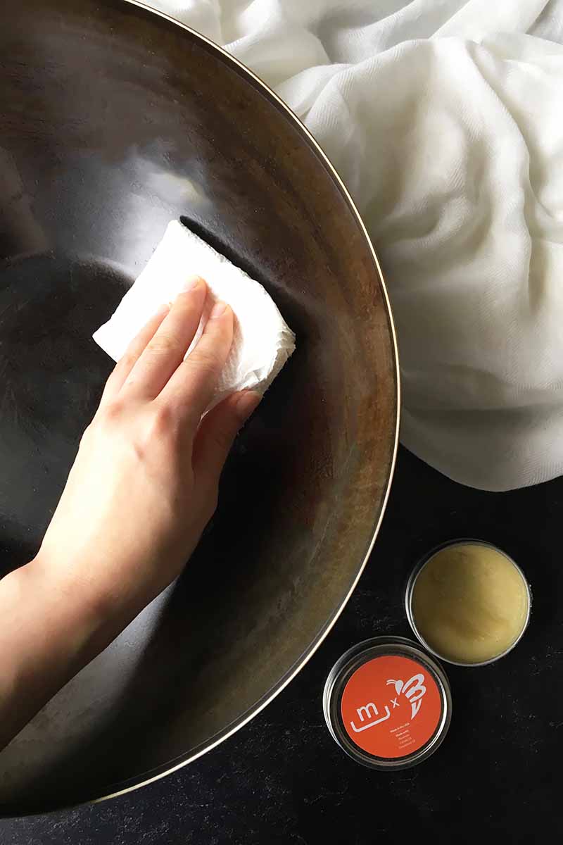 Vertical image of a hand rubbing a pan with a paper towel, with wax on the side.