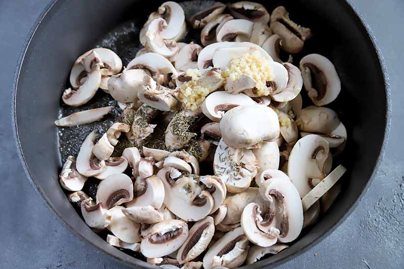 Horizontal image of uncooked sliced button mushrooms and seasonings in a pot.