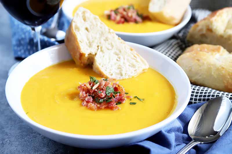 Horizontal image of a white bowl with orange soup, and slices of bread and bacon crumbles.