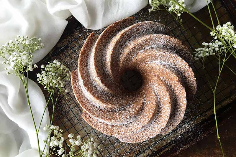 Horizontal top-down image of a whole bundt dusted with sugar on a wire rack, next to white flowers and white towel.