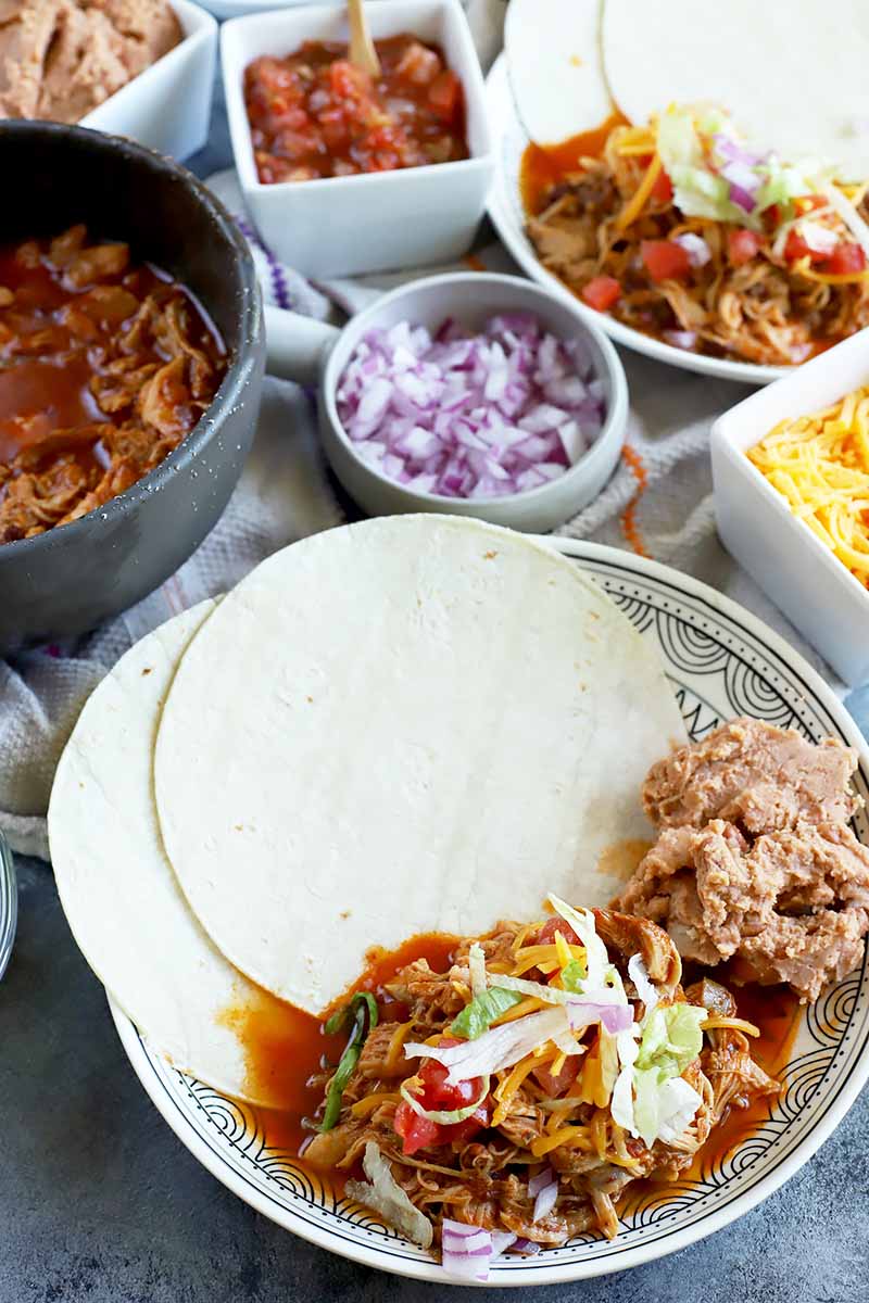 Vertical image of a plate with tortilla wraps, shredded poultry in a red sauce, and various garnishes surrounded by assorted toppings in white dishes.