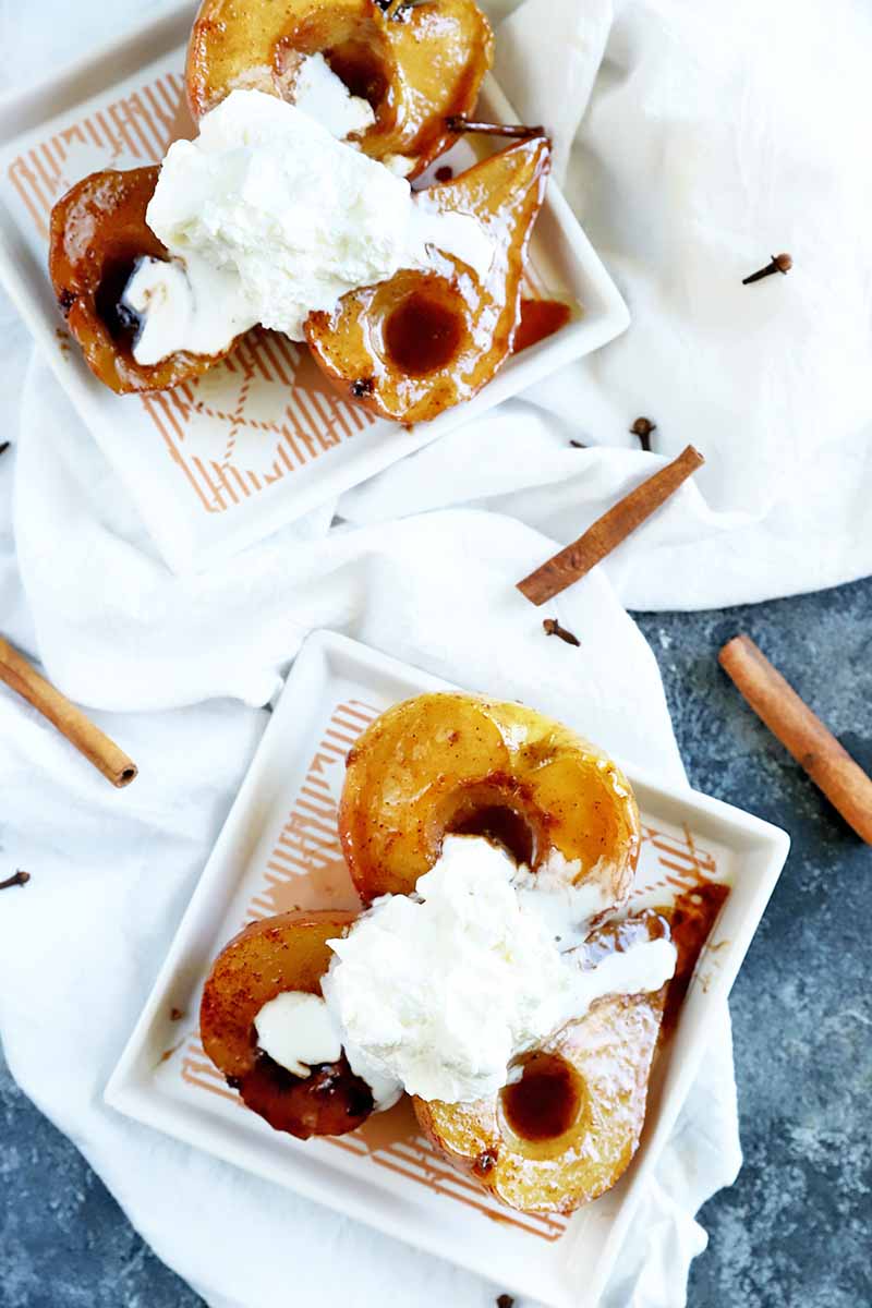 Vertical overhead image of two square white plates of roasted halved pears and apples with caramel sauce, topped with scoops of vanilla ice cream, on a white folded and gathered cloth on top of a gray surface with light and dark speckles, with scattered cinnamon sticks and whole cloves.