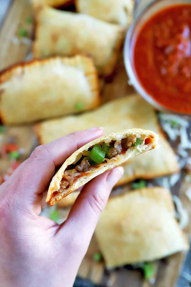 Vertical image of a hand holding half of a pizza pocket to show the sausage and peppers inside, with more of the savory pastries on a cutting board in soft focus in the background, with a small glass bowl of tomato sauce, and scattered cheese and diced vegetables.