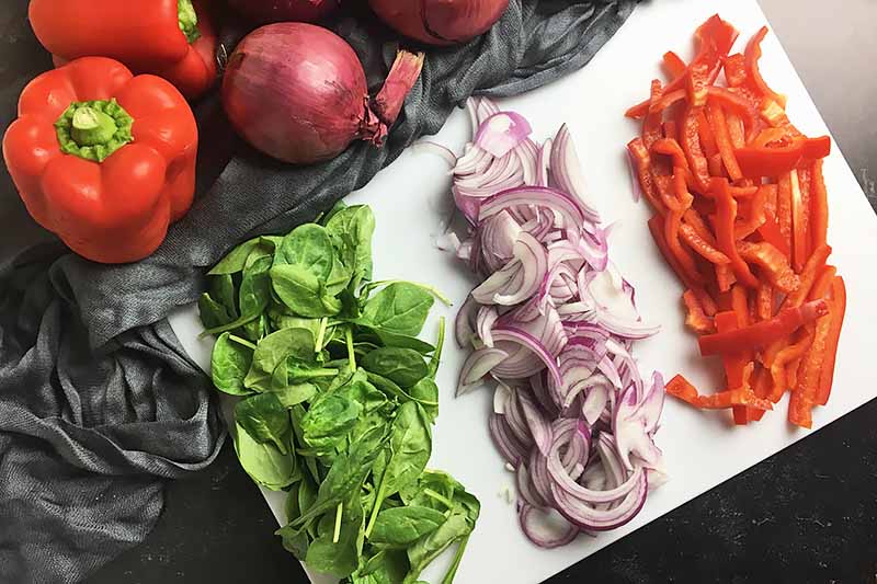 Horizontal image of a cutting board with prepped spinach, red onion, and pepper.
