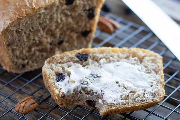 Horizontal image of a slice of pecan and blueberry bread spread with butter on a cooling rack.