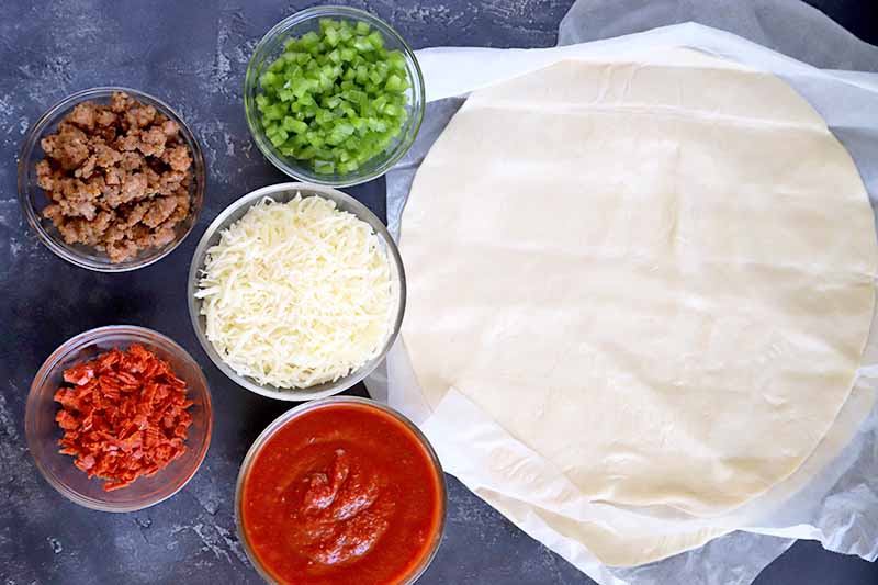 Overhead horizontal image of five small glass bowls of crumbled cooked sausage, chopped sliced pepperoni, diced green bell pepper, shredded mozzarella cheese, and tomato sauce, with several rounds of pizza dough on parchment paper to the right, on a dark blue-gray surface.