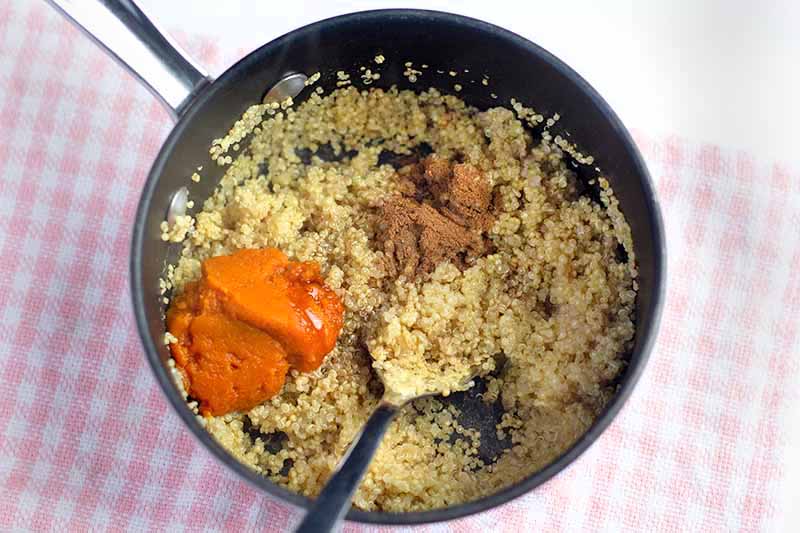 Horizontal overhead image of a nonstick saucepan of cooked quinoa with small piles of pumpkin puree and spices on top, about to be stirred in with a spoon, on a pink and white checkered cloth on top of a white surface.
