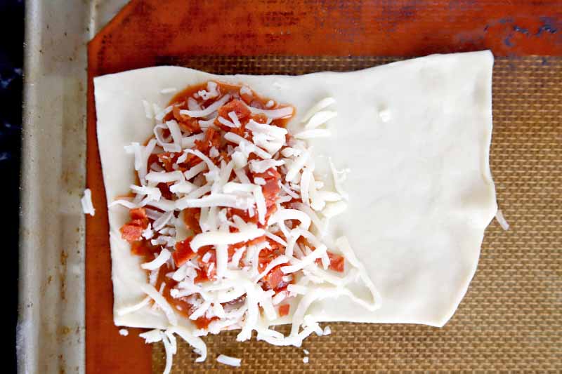 Horizontal overhead closeup image of a rectangle of rolled out dough on a Silpat silicone pan liner resting inside a rimmed metal baking sheet, topped with tomato sauce and shredded mozzarella.