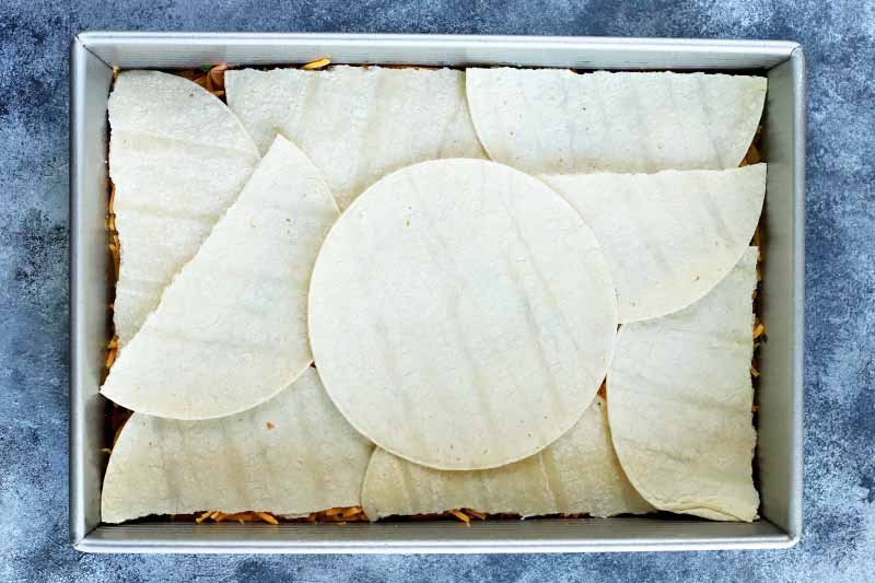 Horizontal overhead image of a rectangular baking pan lined with whole and halved small flour tortillas, on a blue and white mottled and speckled background.