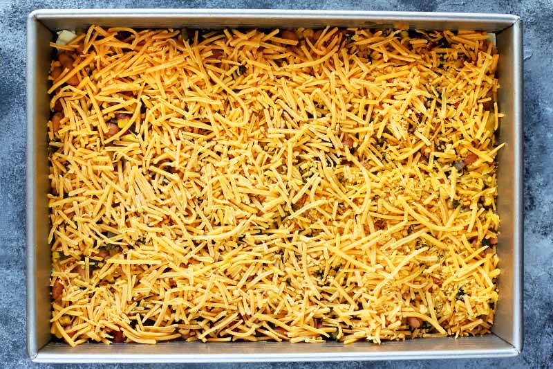 Horizontal overhead image of a metal baking pan filled with a red vegetable mixture topped with orange shredded cheese, on a blue and white surface.