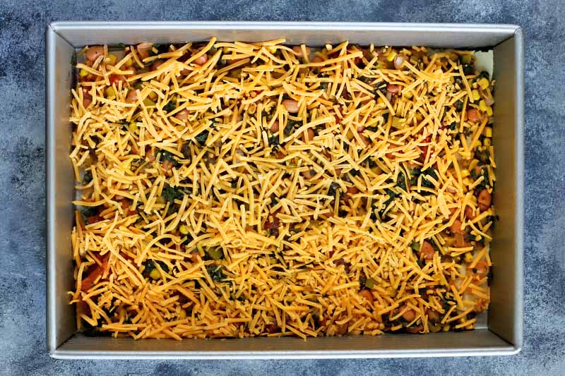 Horizontal overhead image of a rectangular metal baking pan filled with a mixture of vegetables and beans, topped with a layer of shredded cheese, on a blue and white sponge painted surface.