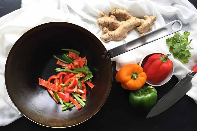 Horizontal image of a pan with assorted peppers next to more peppers, ginger, and a knife on a white towel.
