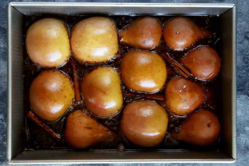 Horizontal overhead image of golden brown roasted halved pears and apples arranged in a rectangular metal baking pan cut side down, surrounded by a brown caramel sauce with whole warming spices, on a gray background.