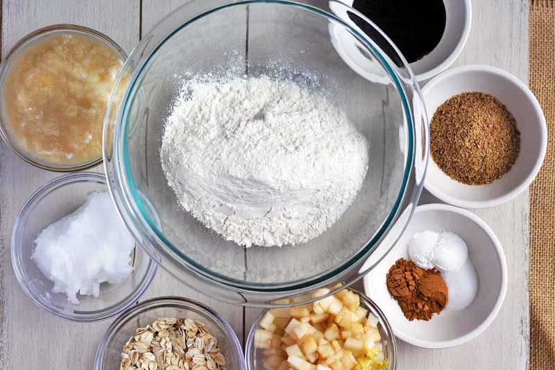 Horizontal overhead image of a clear glass medium-sized mixing bowl of flour, surrounded by smaller glass and white ceramic bowls of molasses, ground flax seed, cinnamon, baking powder, diced pear, applesauce, salt, and baking powder, on a white wood surface.