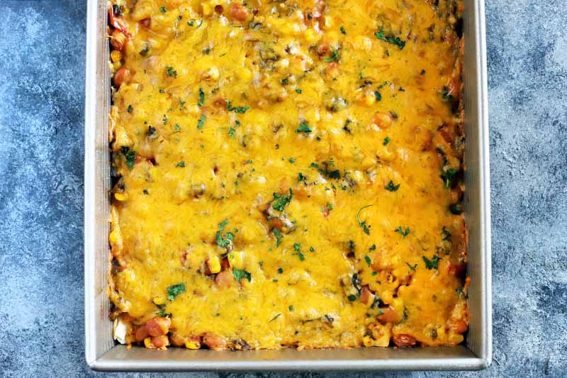 Horizontal overhead image of a closely cropped metal baking pan filled with an enchilada casserole topped with melted cheese, on a blue and white sponge painted surface.