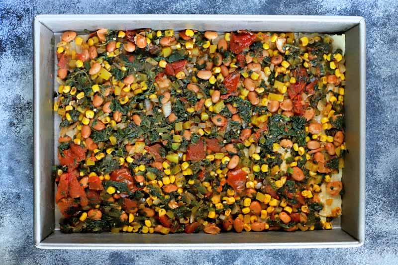 Horizontal overhead image of a metal baking pan filled with an even layer of cooked spinach, tomatoes, beans, and corn, on a blue-gray mottled surface.