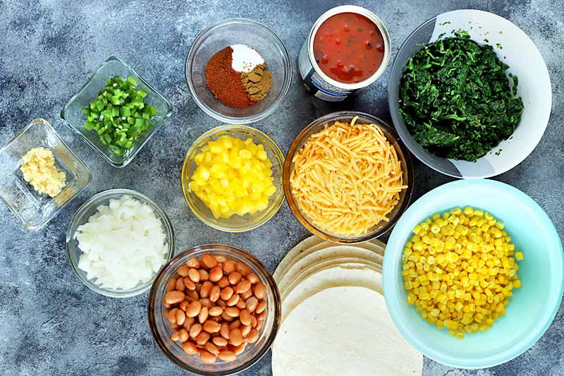 Horizontal overhead image of ceramic glass bowls of various sizes filled with the ingredients required to make a recipe, including chopped jalapeno, chopped onion, chopped yellow bell peppers, minced garlic, beans, spices, a can of tomatoes, spinach, shredded cheese, and corn, with a stack of flour tortillas, on a blue-gray mottled surface.