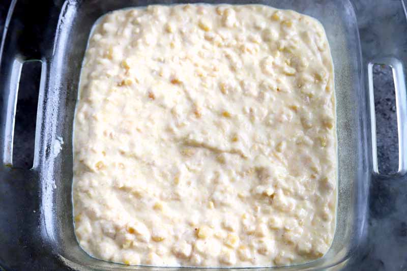Horizontal closely cropped overhead image of a square baking dish with cutout handles, filled with a mixture of creamed corn, sour cream, and baking mix.