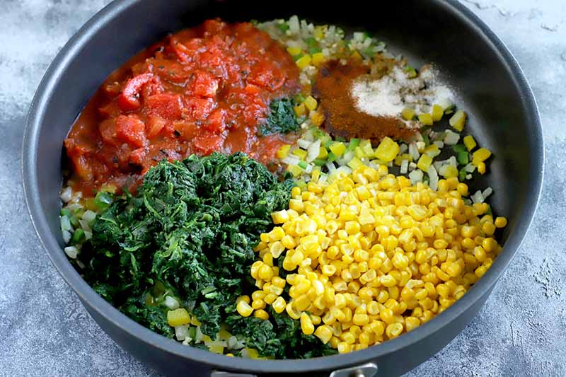 Horizontal image of a large nonstick frying pan filled with separate piles of yellow corn kernels, wilted spinach leaves, canned tomatoes, spiced, and sauteed onion and bell pepper, on a blue and white surface.