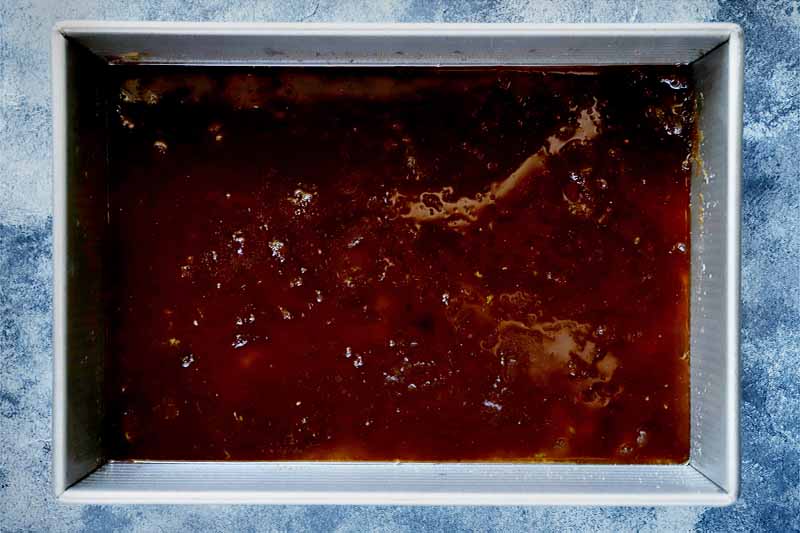 Horizontal overhead image of a brown citrus sauce in a rectangular metal baking pan, on a blue-gray speckled background.