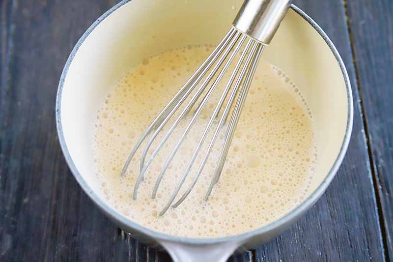 Horizontal image of a whisk in a pot with a frothy, milky liquid.