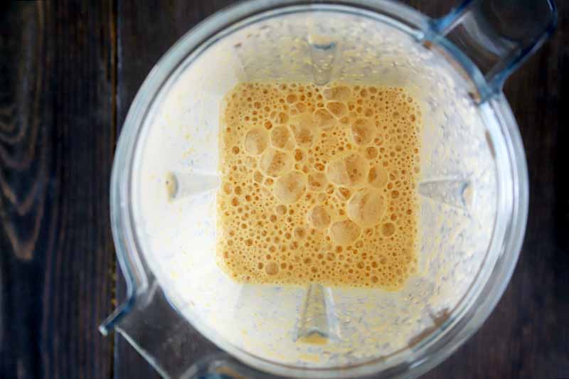 Horizontal overhead image of a frothy orange blended pumpkin smoothie in the bottom of a clear plastic blender pitcher, on a dark brown wood surface.