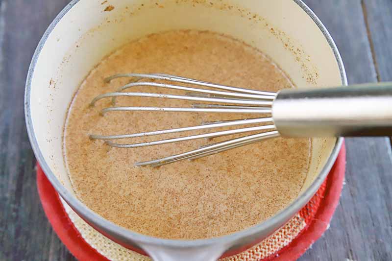 Horizontal image of a whisk mixing a liquid and spice mixture in a pot.