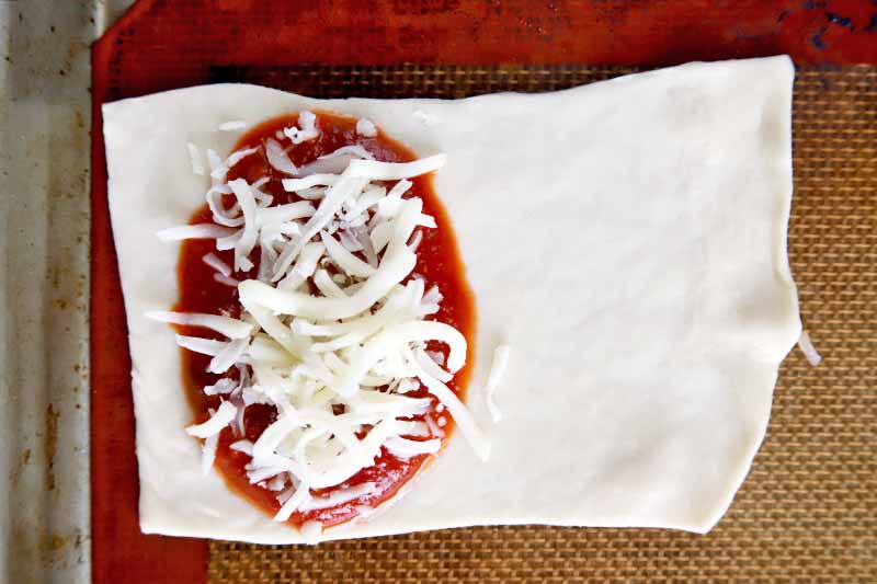 Overhead horizontal image of a rectangular rolled out portion of dough topped with red sauce and shredded cheese, on an orange and beige Silpat silicone liner set into a rimmed metal baking sheet.