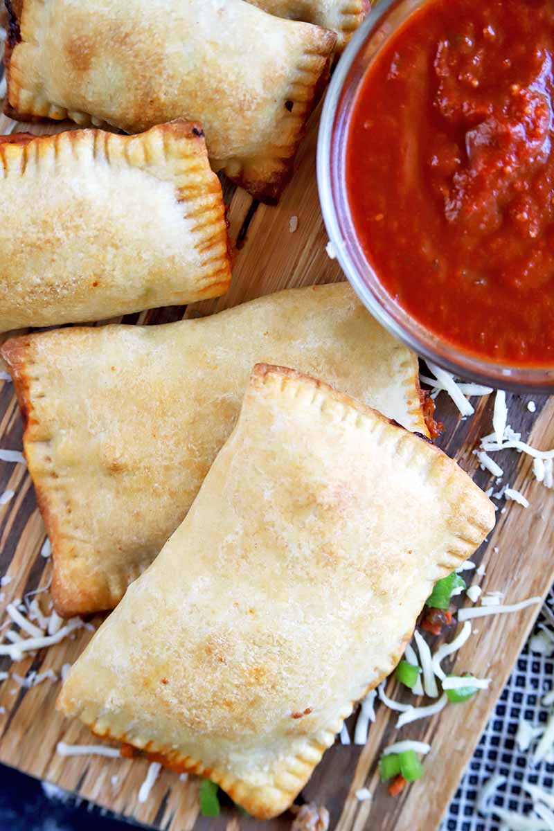 Vertical overhead image of four rectangular pizza pocket pastries on a wooden cutting board, with a glass bowl of tomato sauce and scattered cheese shreds and diced green bell pepper, on a cloth surface.