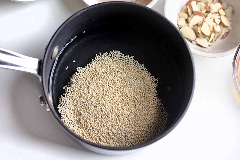 Horizontal overhead image of a nonstick saucepan of uncooked quinoa and water, on a white surface with a small white dish of sliced almonds.