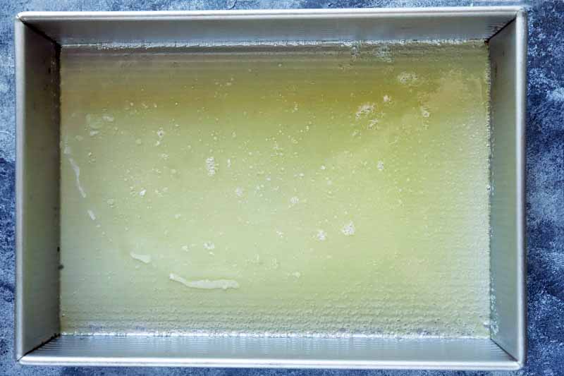 Horizontal overhead image of melted butter in a rectangular metal baking pan, on a blue-gray sponge painted background.