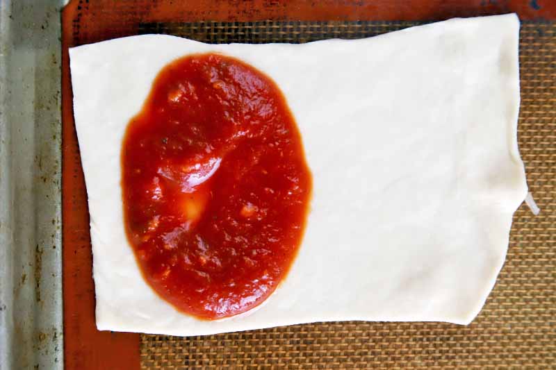 Horizontal overhead image of a rectangular portion of rolled out dough topped with red sauce, on an orange and beige Silpat liner set into a rimmed metal baking sheet pan.