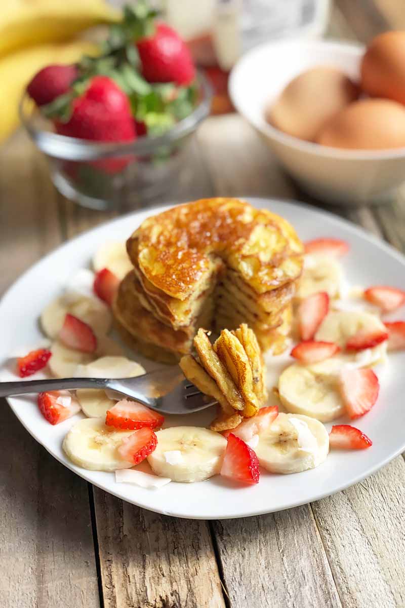 Vertical image of a fork with a stack of golden brown pancakes on a white platter with sliced fruit.