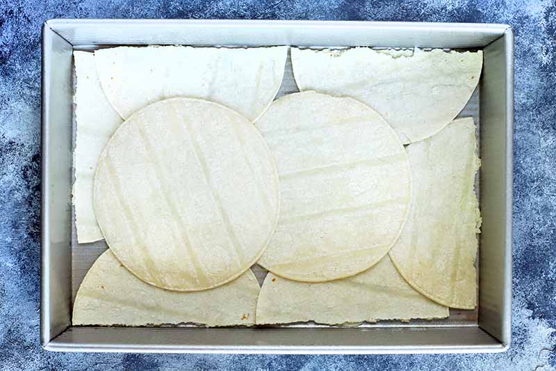 Horizontal overhead image of a metal rectangular baking pan lined with flour tortillas, some whole and some torn in half, on a blue and white painted surface.