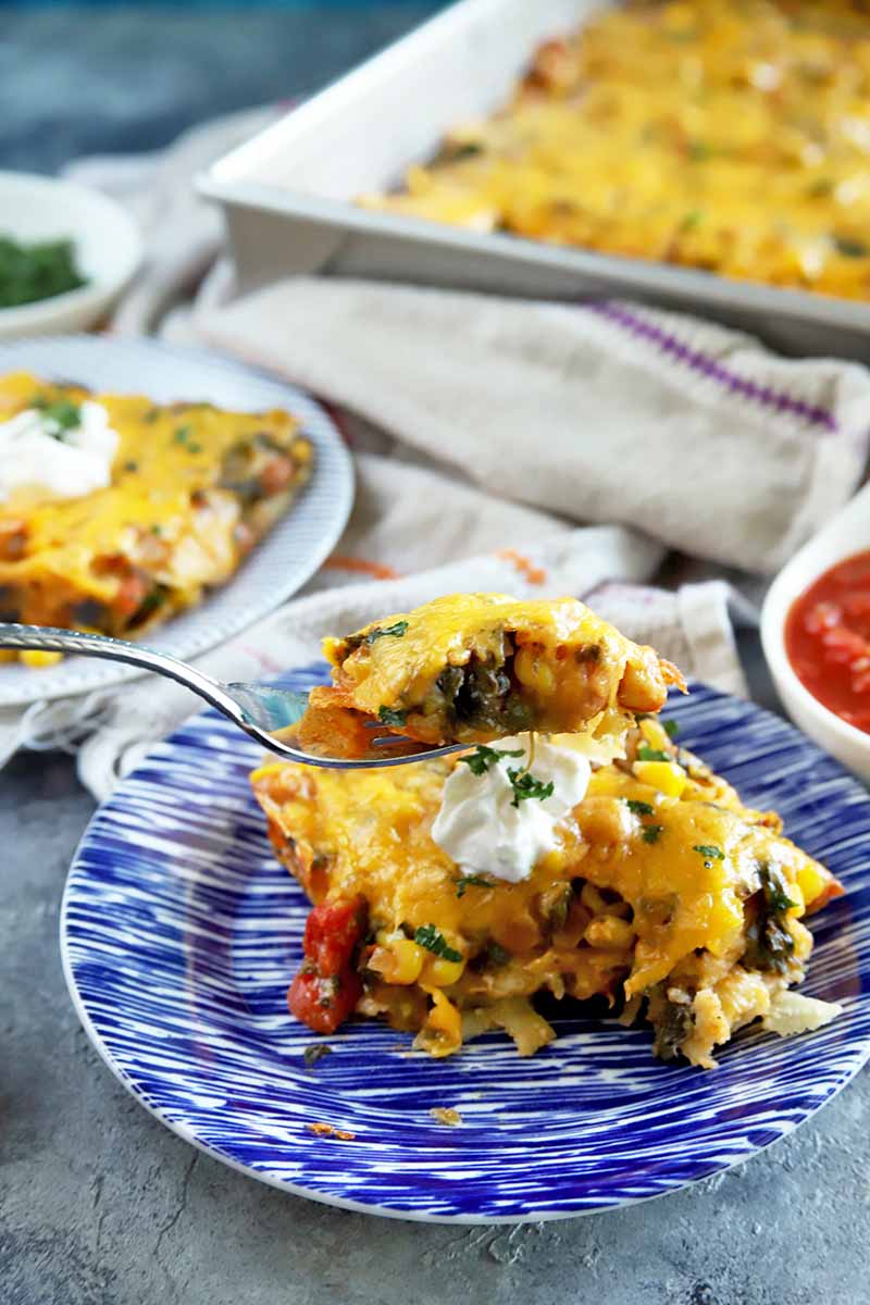 Vertical image of a forkful of stacked enchilada casserole being held up to the camera, with more of the disho n blue and white plates in the background and in a metal baking pan, with a white dish towel and small white ceramic dishes of chopped fresh herbs and red salsa.