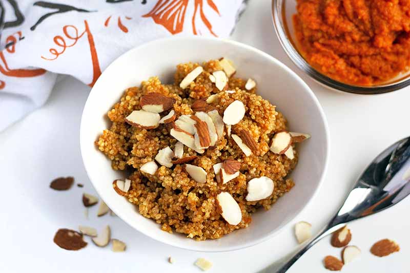 Overhead horizontal image of a white bowl of quinoa breakfast cereal with pumpkin topped with sliced almonds, on a white surface with scattered nuts, an autumn-themed dish towel, a bowl of orange roasted squash puree, and a spoon.