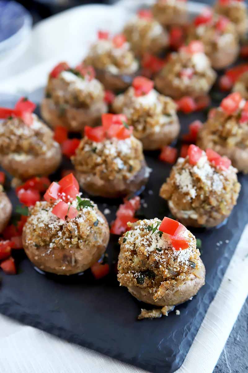 Vertical image of rows of stuffed mushrooms with tomato garnish on a black slate.