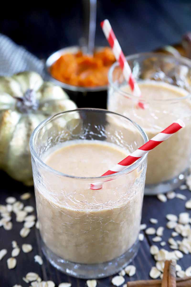 Vertical image of two glasses of homemade pumpkin oatmeal smoothie with red and white striped paper straws, a decorative metallic gold pumpkin, a small dish of orange squash puree, and scattered uncooked oats and cinnamon sticks, on a dark gray surface against a black background.