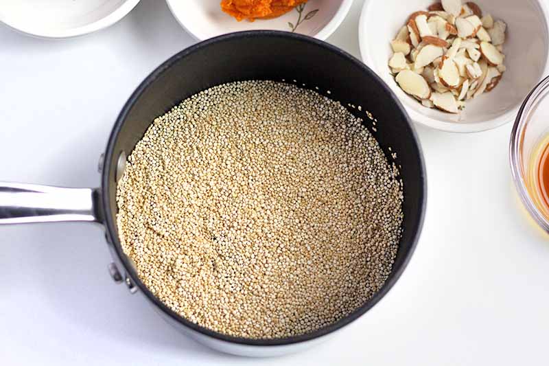 Horizontal overhead image of a nonstick saucepan of quinoa, surrounded by small white bowls of pureed pumpkin, sliced almonds, and other ingredients, on a white countertop.