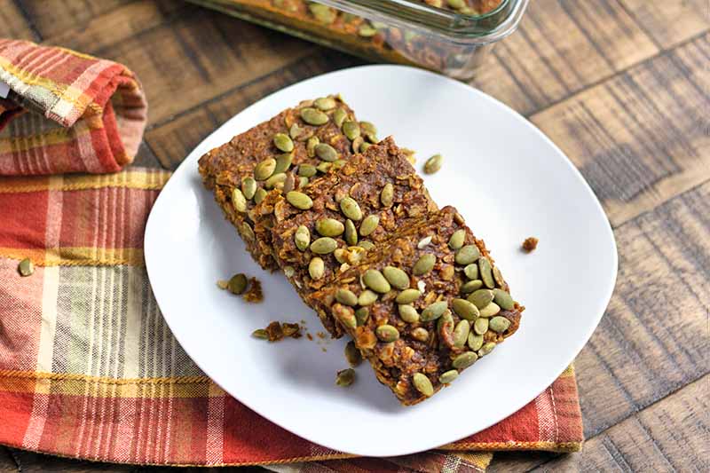 Horizontal image of shingled pumpkin squares topped with pumpkin seeds on a white plate with a plaid towel.