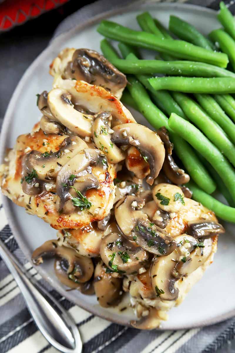 Vertical top-down image of pan-seared chicken topped with mushrooms next to green beans on a plate.