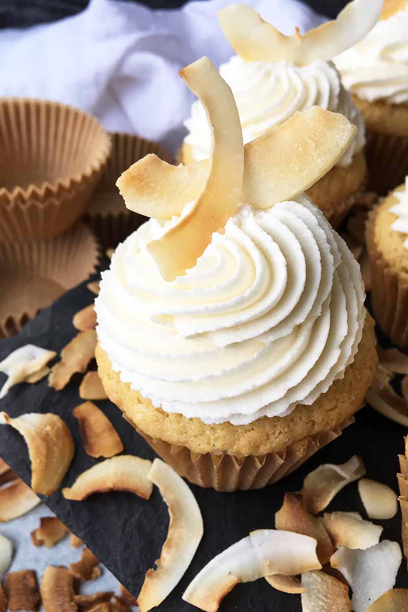 Vertical image of vanilla baked goods topped with white frosting and golden-brown garnishes in front of empty muffin liners.