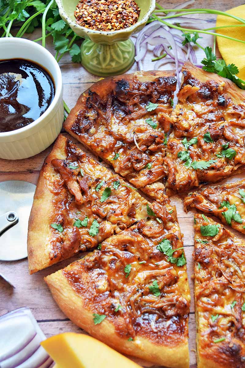 Vertical overhead closely cropped image of a barbecue chicken pizza on a wood table with chunks of gouda, cups of red pepper flakes and barbecue sauce, sprigs of fresh herbs, and a yellow cloth.