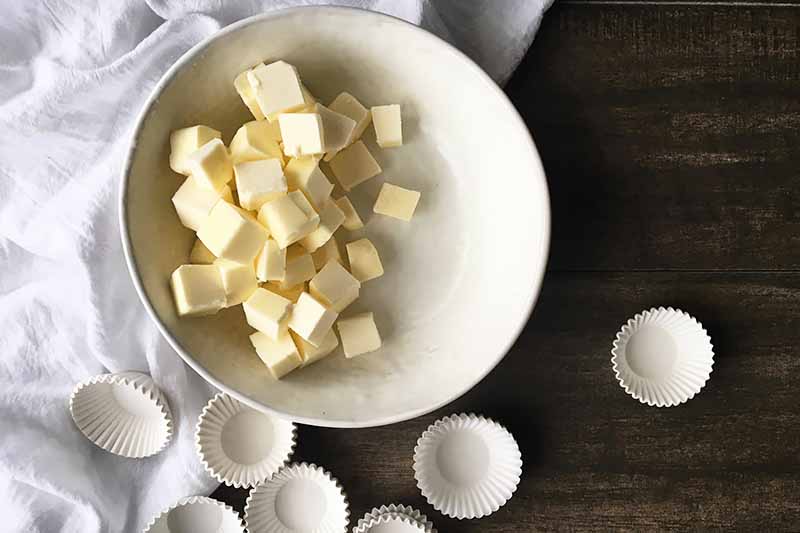 Horizontal image of a white bowl with cubes of butter next to mini cupcakes on a dark wooden board.