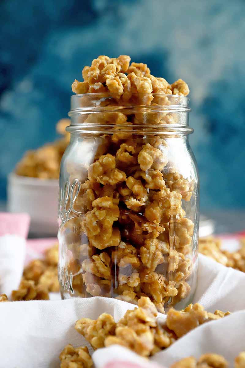Vertical head-on image of a glass jar overflowing with homemade caramel corn, with more scattered on a folded and gathered white cloth, with a white bowl of the snack in soft focus in the background, against a mottled blue and white backdrop.