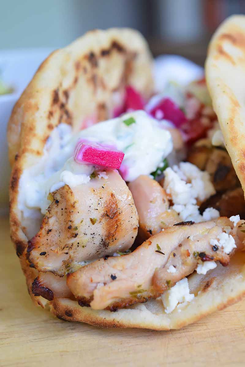Vertical image of grilled marinated chicken thigh meat on a piece of pita with pickled vegetables and yogurt sauce.
