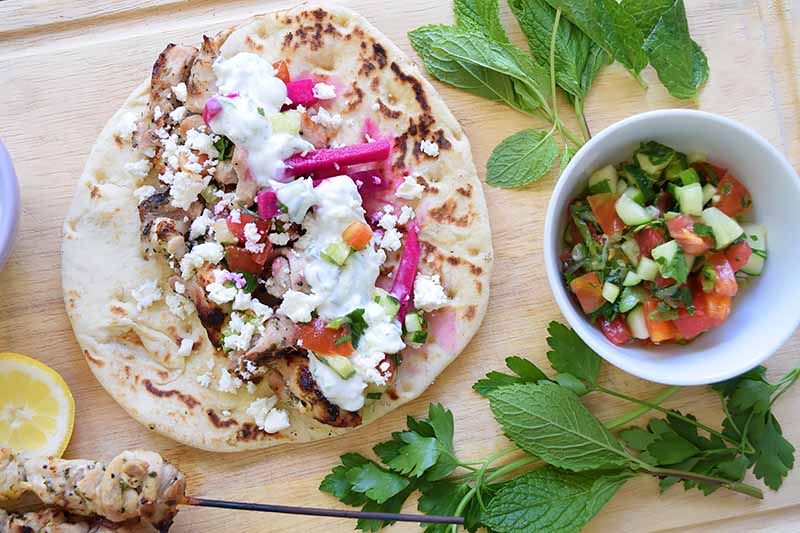 Horizontal overhead image of an open-faced chicken gyro with various vegetables, feta cheese, and tzatziki, on a wooden cutting board with sliced lemon, a bamboo skewer of grilled poultry, sprigs of fresh Italian flat leaf parsley and mint, and a small white bowl of tomato salad with cucumbers.
