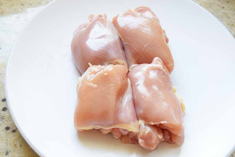Horizontal overhead image of boneless skinless chicken thighs on a white plate, on an off-white speckled surface.